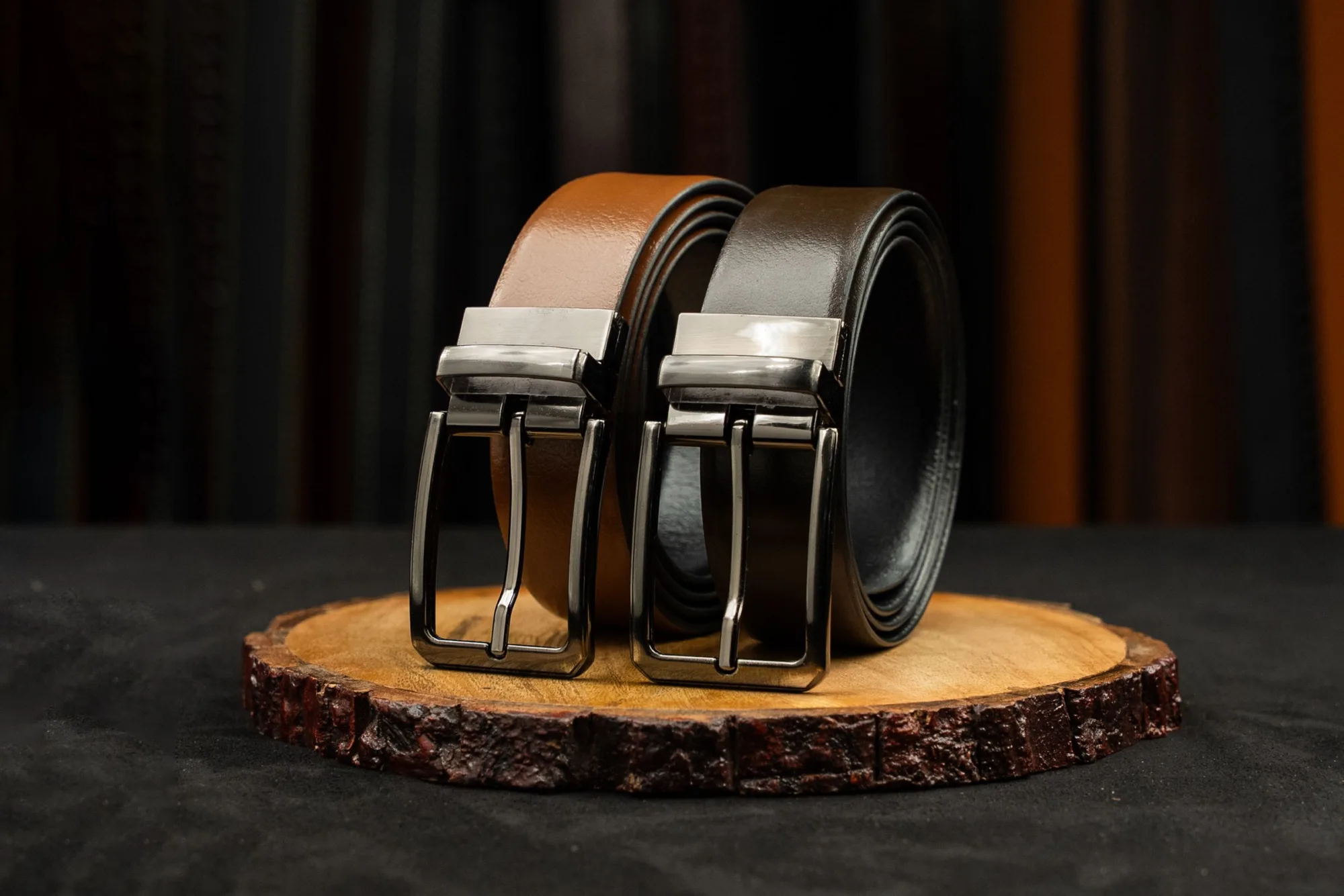 Two leather belts with metal buckles displayed on a rustic wooden platform