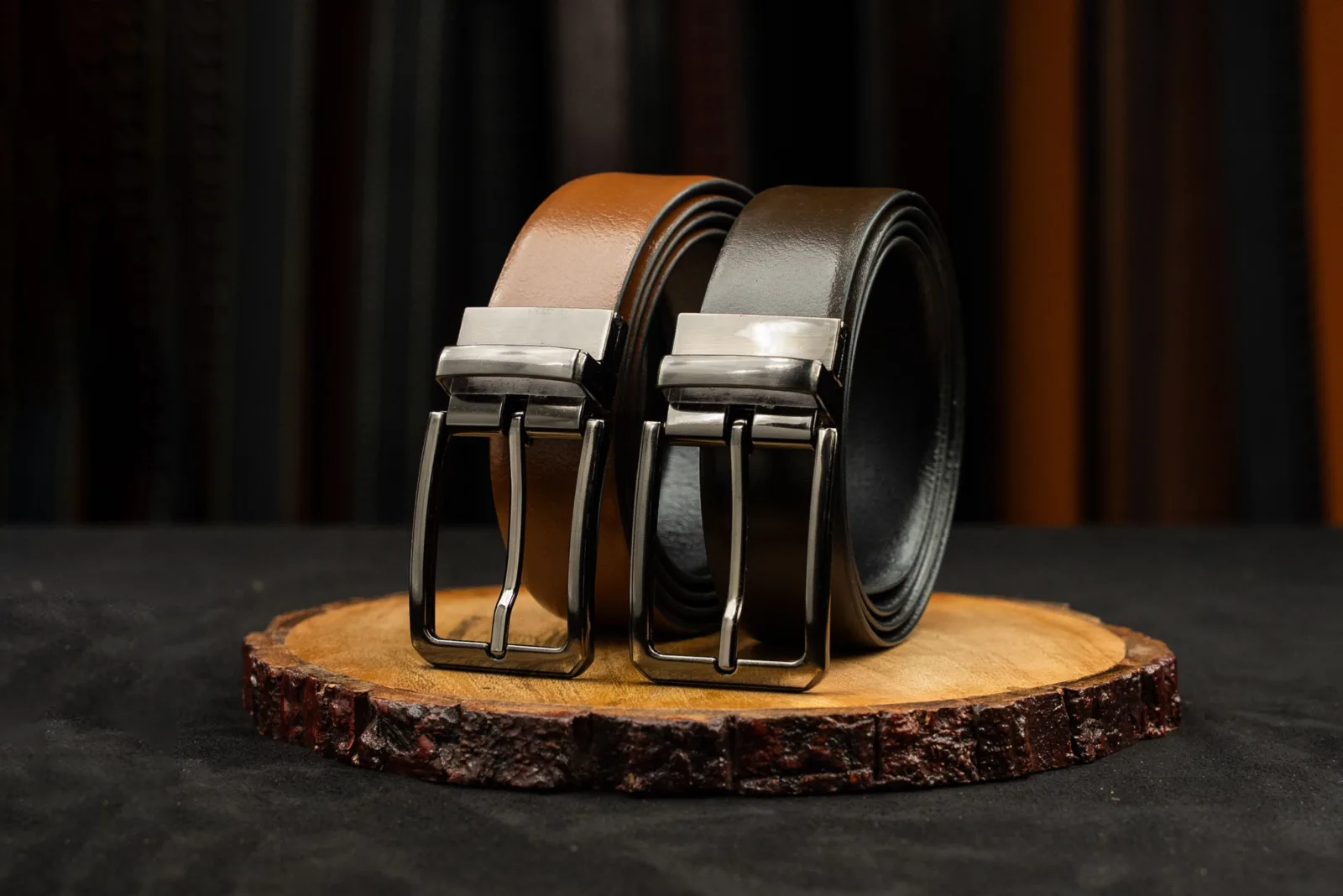 Two leather belts with metal buckles displayed on a rustic wooden platform